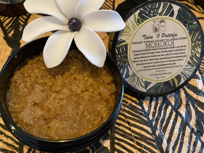 K&ouml;nnte beinhalten: Ein schwarzer Topf gef&uuml;llt mit einem braunen Zuckerschrub. Der Topf hat eine wei&szlig;e Blume oben und ein Etikett mit der Aufschrift "Tiare O Patitifa Organic Tongan Coconut Oil Sugar Scrub MOHOKOI".