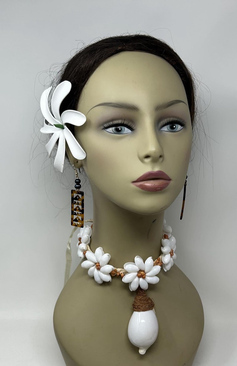May include: A white flower hair clip and a white shell necklace with a brown cord and a large white shell pendant. The necklace is on a mannequin with brown hair.
