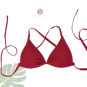 May include: A solid red bikini top with a triangle design and crisscross straps. The top has long, thin ties for adjusting the fit. The background is white with a green leaf in the lower left corner.