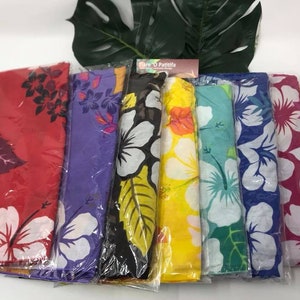 May include: A collection of colorful sarongs, each wrapped in clear plastic. The sarongs feature floral patterns with hibiscus flowers in various colors, including red, purple, black, yellow, teal, blue, pink, and orange. A green leaf is visible in the background.