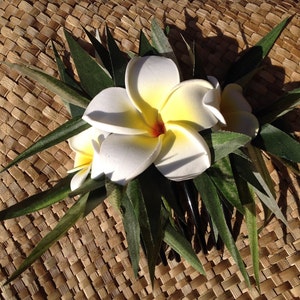 May include: A white and yellow flower hair comb with green leaves. The comb is on a woven brown background.