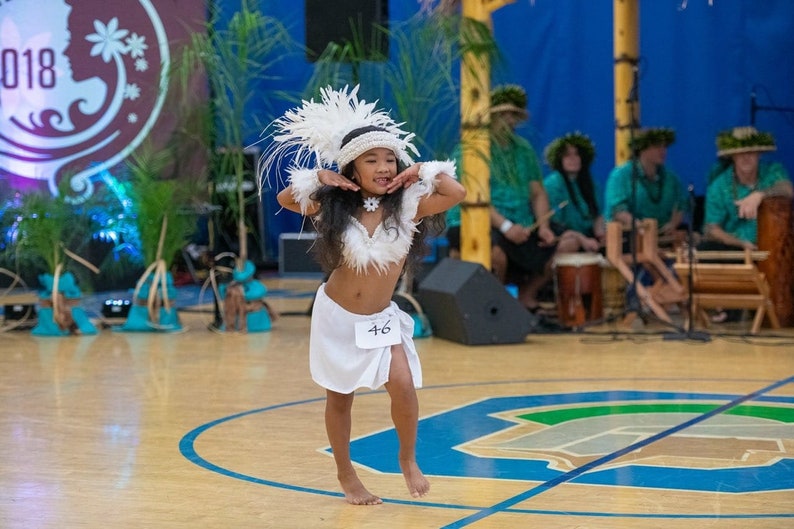 May include: A young girl in a white costume with feathers and a white skirt dances on a wooden floor. She is wearing a white flower necklace and has a number 46 on her skirt.