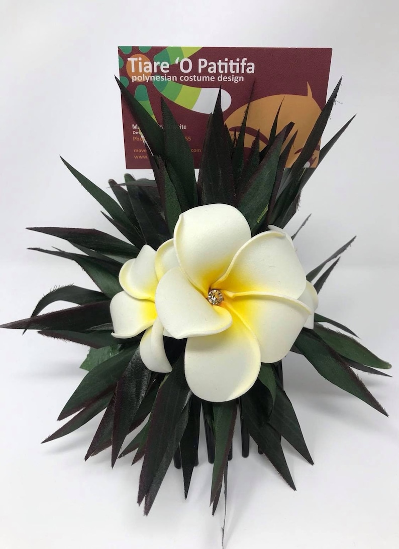 May include: A white and yellow plumeria flower hair comb with green leaves. The comb is decorated with a small rhinestone. The comb is labeled "Tiare 'O Patitfa Polynesian costume design".
