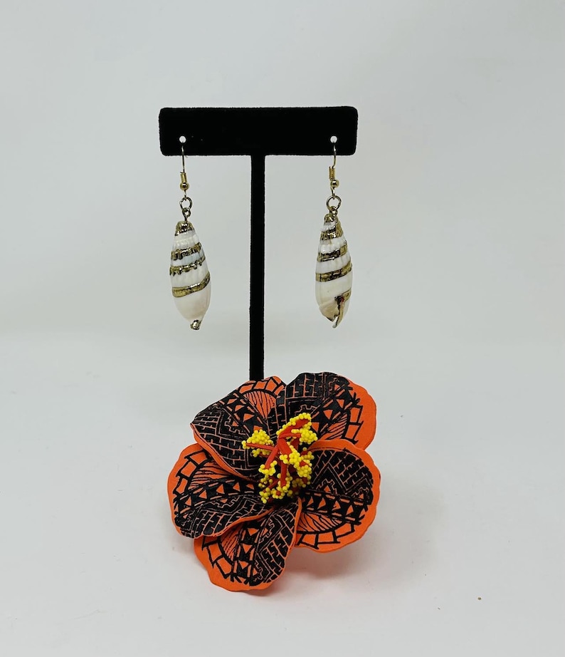 May include: A pair of white and gold shell earrings hanging on a black stand with a red and black hibiscus flower below.