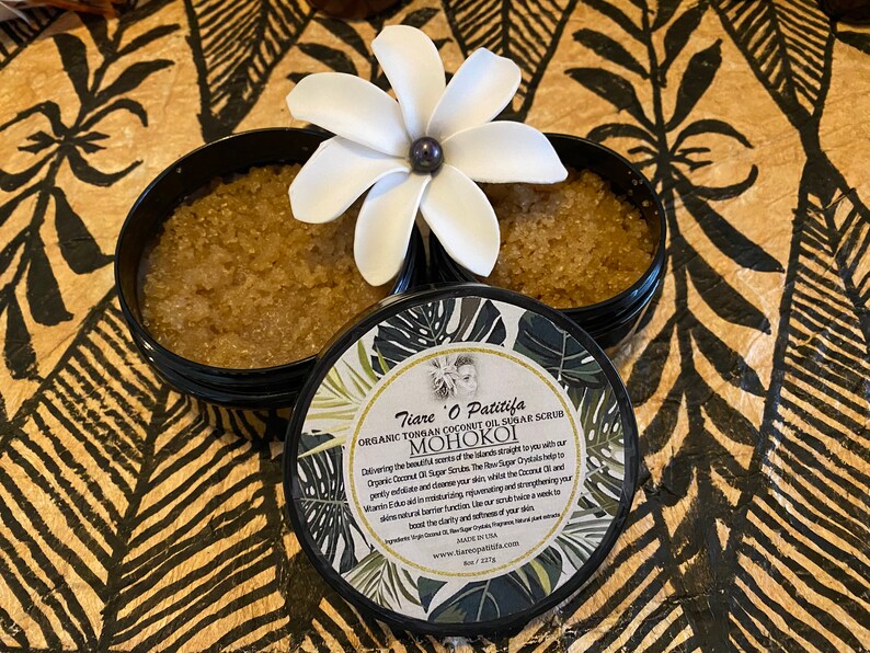 K&ouml;nnte beinhalten: Ein schwarzer Topf mit einem wei&szlig;en Deckel, der ein braunes Zuckerschrub enth&auml;lt. Der Topf hat ein Etikett mit dem Text "Tiare O Patitifa Organic Tongan Coconut Oil Sugar Scrub MOHOKOI Delivering the beautiful scents of the Islands straight to you with our Organic Coconut Oil and Sugar Scrub. The Raw Sugar Crystals help to gently exfoliate and cleanse your skin, whilst the Coconut Oil and Vitamin E work in barrier function, rejuvenating and strengthening your skin's natural moisturising factors.  Our scrub helps to provide a warm, exotic add to the clarity and softness of your skin.  Made with the best of Tongan and Hawaiian natural plant ingredients.  Made in the USA. www.tiareopatitifa.com Net Wt 227g"