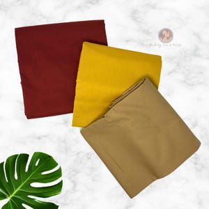 May include: Three folded pieces of fabric in solid colors: one deep burgundy, one mustard yellow, and one tan. The fabric pieces are arranged in a stack, with a green monstera leaf visible in the lower left corner of the frame.