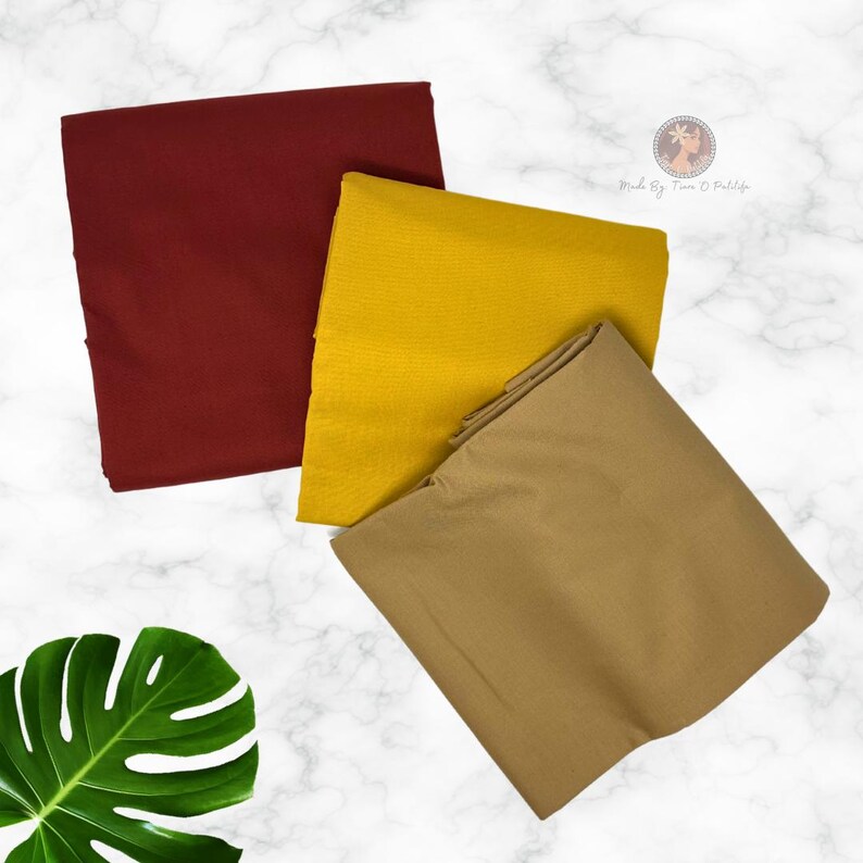 May include: Three folded pieces of fabric in solid colors: one deep burgundy, one mustard yellow, and one tan. The fabric pieces are arranged in a stack, with a green monstera leaf visible in the lower left corner of the frame.
