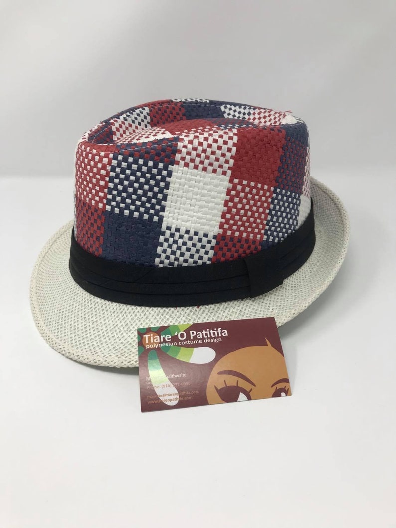 May include: A white and red checkered fedora hat with a black band. The hat is made of woven straw and has a wide brim. The hat is sitting on a white surface. The hat has a label that says "Tiare 'O Patitifa Polynesian costume design".