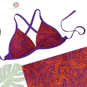 May include: A two-piece bikini set featuring a vibrant orange and purple tribal pattern. The bikini top has a triangle shape with purple trim and straps. The matching bottom is partially visible, showcasing the same intricate design. The set is made by Tiana & Co.