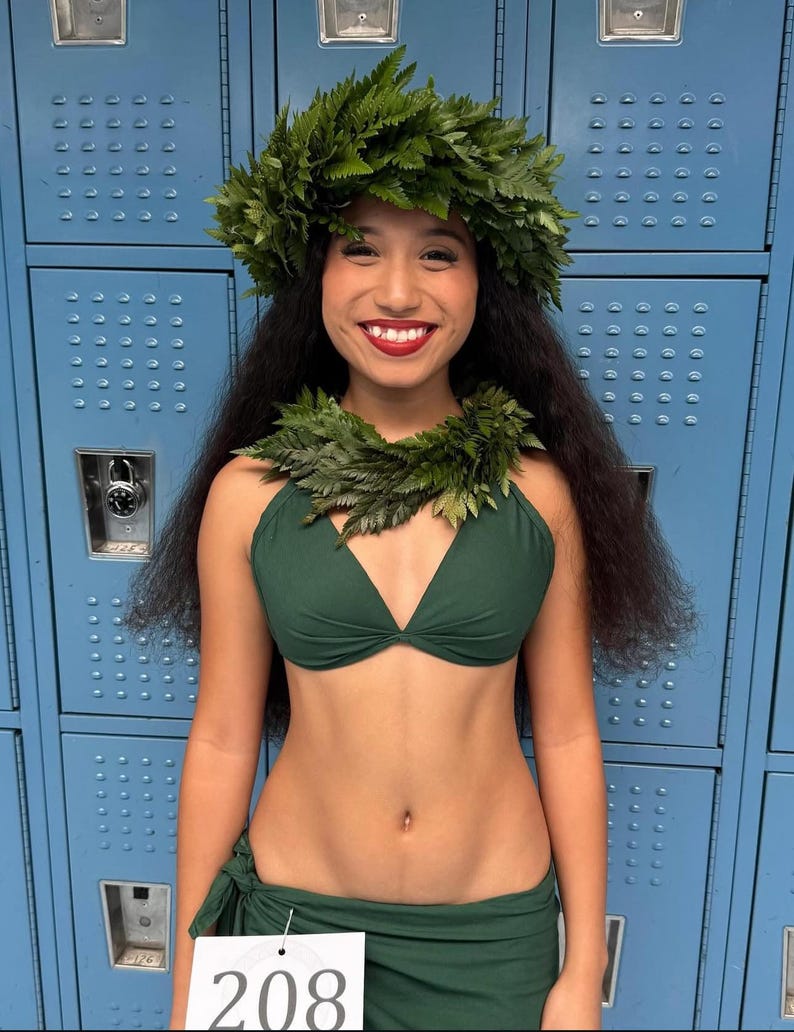 May include: A dark green two-piece outfit consisting of a halter-style bikini top and a matching wrap skirt. The ensemble is paired with a vibrant green fern leaf headpiece and a matching fern leaf lei worn around the neck. A white tag with the number 208.