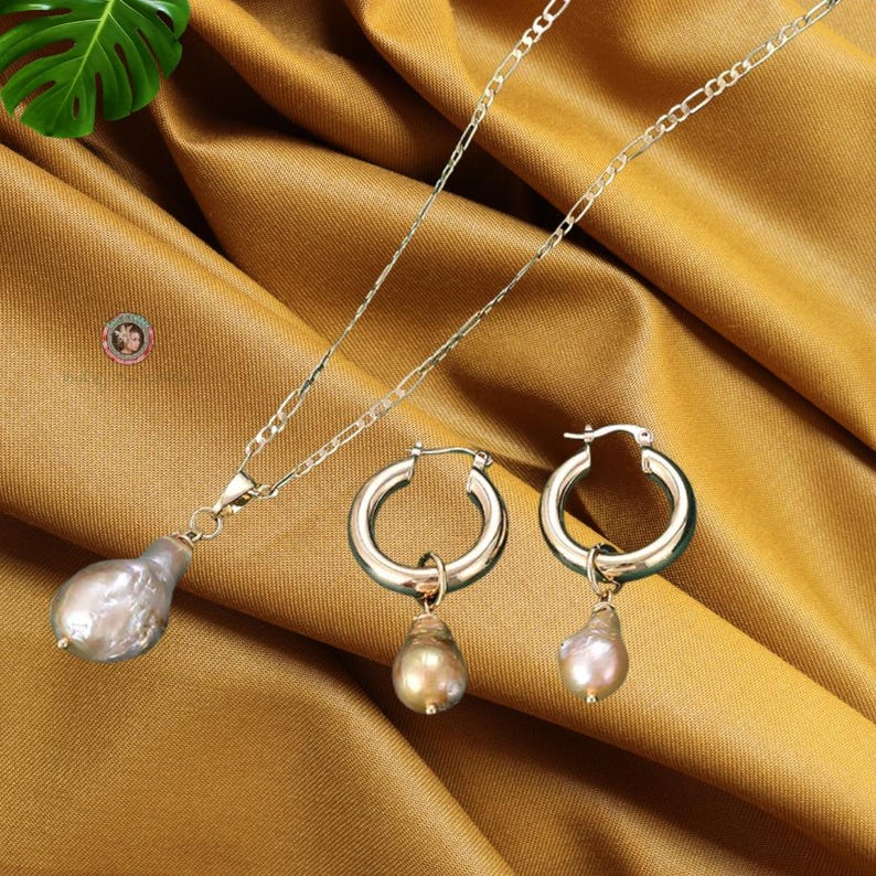 May include: A gold chain necklace with a large, pearlescent, gray and white pearl pendant. Matching hoop earrings with a smaller, pearlescent, gray and white pearl pendant.