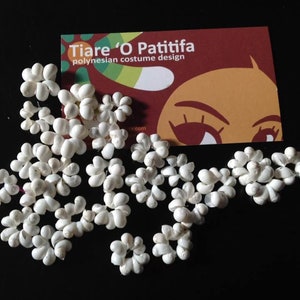 May include: A close-up of a white flower garland made of small, white, flower-shaped pieces. The garland is arranged in a circular pattern and is lying on a black surface. The garland is a traditional Polynesian costume design.