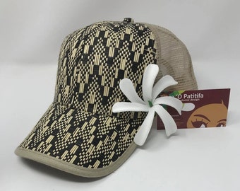 May include: A beige and black patterned baseball cap with a mesh back. A white flower is attached to the side of the cap. The cap is sitting on a white surface. The text "O Patitifa Costume design" is visible on a business card in the background.