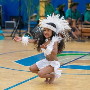 May include: A young girl in a white feathered headdress and skirt performs a traditional dance on a wooden floor. She is wearing a white flower necklace and has her hair pulled back.