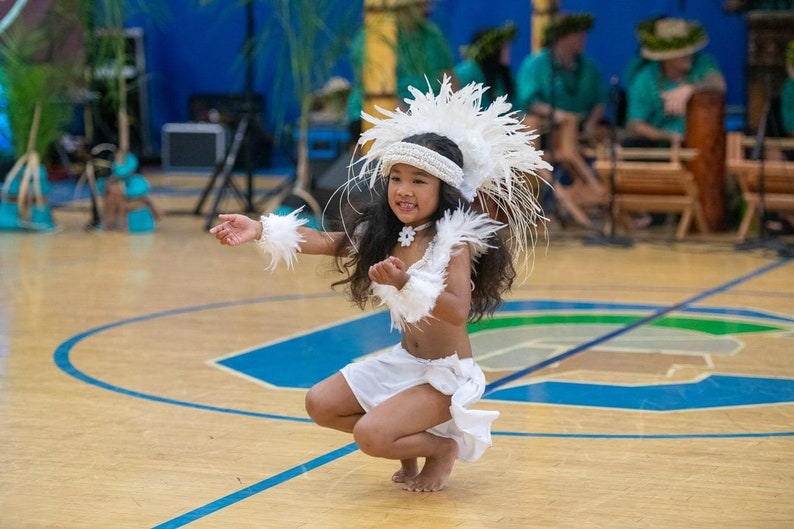 May include: A young girl in a white feathered headdress and skirt performs a traditional dance on a wooden floor. She is wearing a white flower necklace and has her hair pulled back.