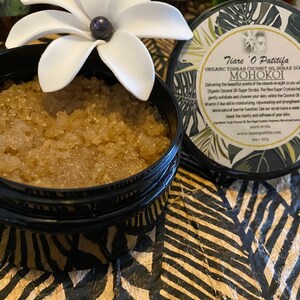 K&ouml;nnte beinhalten: Ein schwarzer Topf mit einer wei&szlig;en Blume oben drauf, gef&uuml;llt mit einem braunen Zuckerschrub. Der Topf hat ein Etikett mit der Aufschrift "Tiare O Patitifa Organic Tongan Coconut Oil Sugar Scrub MOHOKOI".