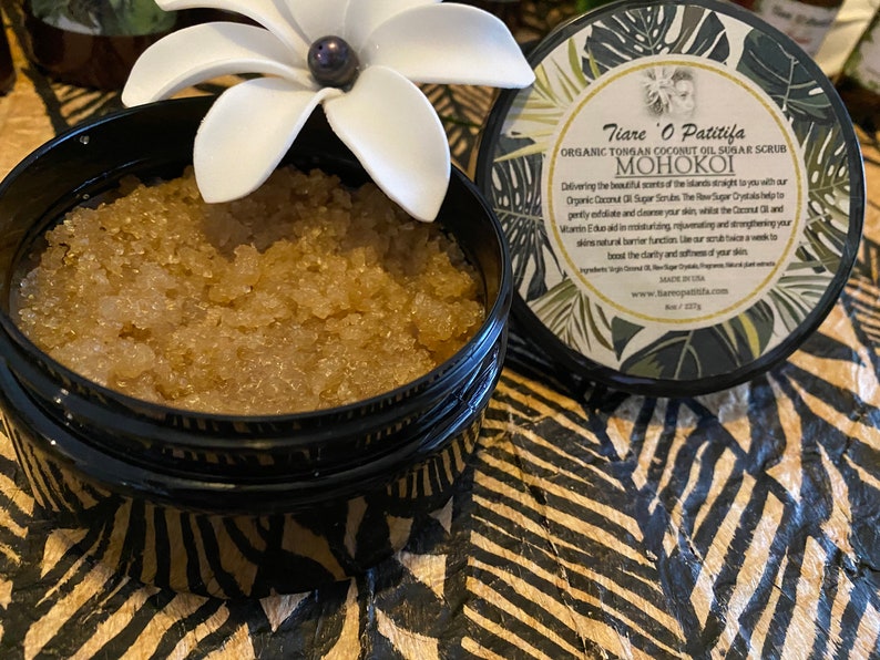 K&ouml;nnte beinhalten: Ein schwarzer Topf mit einer wei&szlig;en Blume oben drauf, gef&uuml;llt mit einem braunen Zuckerschrub. Der Topf hat ein Etikett mit der Aufschrift "Tiare O Patitifa Organic Tongan Coconut Oil Sugar Scrub MOHOKOI".