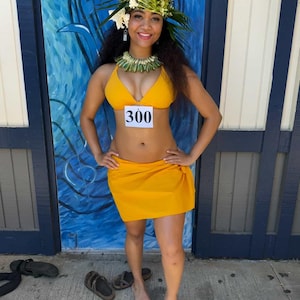 May include: A bright yellow two-piece bikini set consisting of a triangle top and a wrap-style skirt. The top features a white rectangular tag with the black number 300 printed on it. The outfit is accessorized with a green leaf headpiece and necklace.