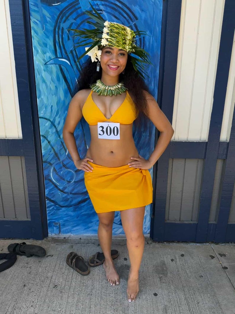 May include: A bright yellow two-piece bikini set consisting of a triangle top and a wrap-style skirt. The top features a white rectangular tag with the black number 300 printed on it. The outfit is accessorized with a green leaf headpiece and necklace.