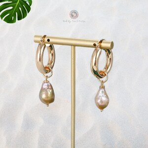 May include: A pair of gold hoop earrings with a pearlescent teardrop charm hanging from each hoop. The earrings are displayed on a gold stand.