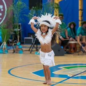 May include: A young girl in a white feathered costume dances on a wooden floor. She is wearing a white feather headdress, a white feather top, and a white skirt. She has a number 46 on her skirt. She is smiling and looking at the camera.