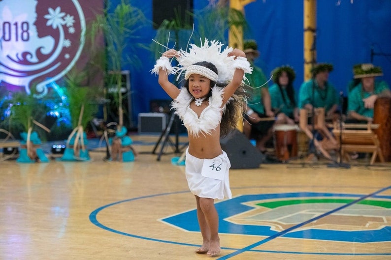 May include: A young girl in a white feathered costume dances on a wooden floor. She is wearing a white feather headdress, a white feather top, and a white skirt. She has a number 46 on her skirt. She is smiling and looking at the camera.