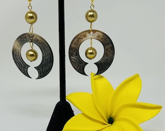 May include: A pair of gold earrings with a large, circular, carved, brown and black pendant with a gold bead hanging from the bottom. The earrings are hanging from a black stand with a yellow flower in the foreground.