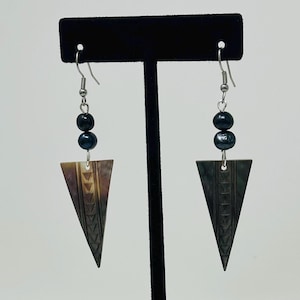 Puede incluir: Un par de pendientes de plata con detalles de perlas negras y un colgante de forma triangular con un patrón geométrico.