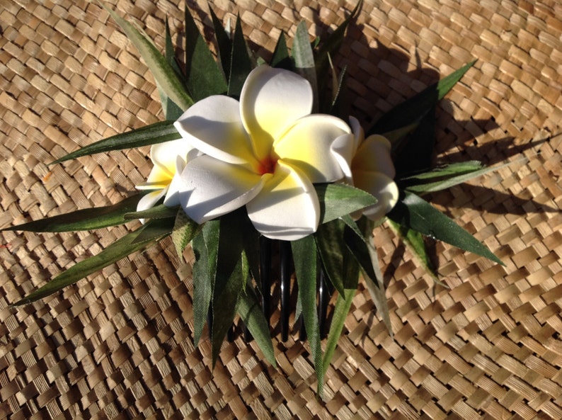 May include: A white and yellow flower hair comb with green leaves. The comb is on a woven brown background.