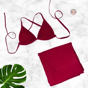 Könnte beinhalten: Ein zweiteiliges bordeauxfarbenes Badeanzug-Set. Das Oberteil ist ein Triangel-Bikini mit verstellbaren Trägern. Das Unterteil ist ein hoch taillierter Slip. Das Set wird auf einer weiß-grauen Marmoroberfläche präsentiert. Ein großes grünes Blatt befindet sich in der linken unteren Ecke.