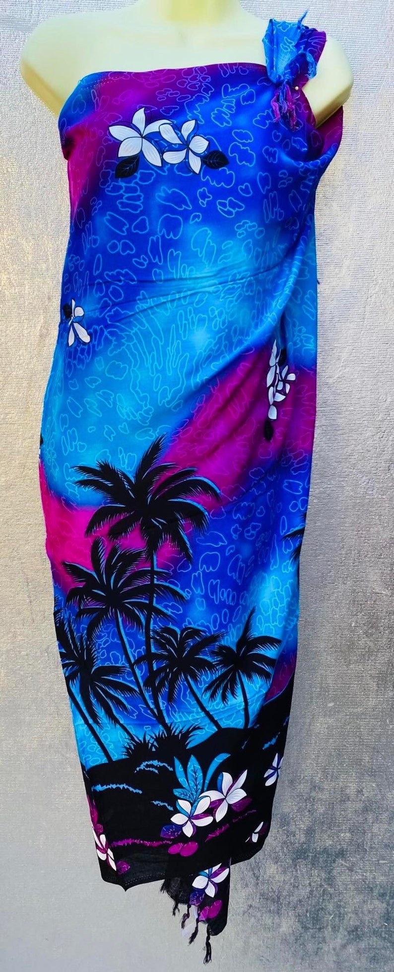 Puede incluir: Un sarong azul y morado con un estampado tropical de palmeras y flores blancas. El sarong tiene un borde con flecos.
