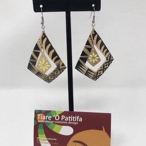 May include: Geometric wooden earrings with black and tan tribal patterns and a yellow flower design. The earrings are displayed on a black stand. A business card with the text "Tiare 'O Patitifa" is in the foreground.