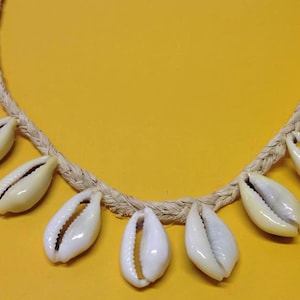 May include: A necklace made of natural fiber cord with white cowrie shells hanging from it. The necklace is on a yellow background.
