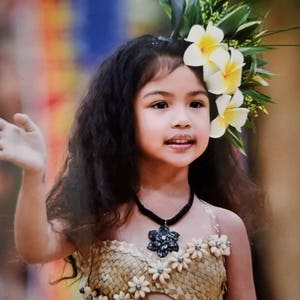 May include: A young girl with long dark curly hair wears a woven flower-shaped top with a black flower pendant necklace. She is wearing a flower crown with white plumeria flowers and green leaves.