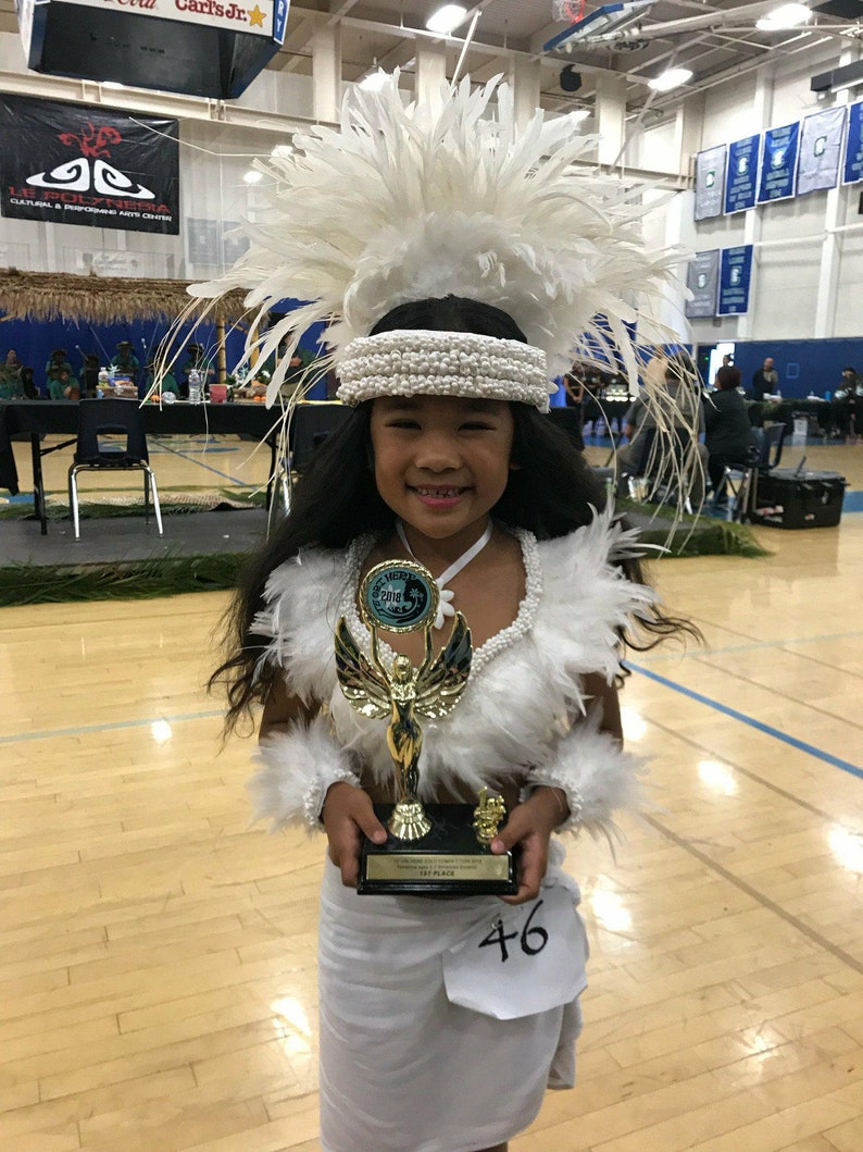 May include: A young girl wearing a white feather headdress and a white dress holds a gold trophy with a blue ribbon. The trophy has a golden angel figure on top. The girl is smiling and has a number 46 on her dress.