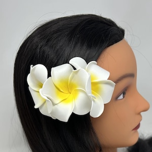 Puede incluir: Una pinza para el pelo con una flor de plumeria blanca y amarilla. La flor tiene pétalos blancos con centros amarillos. El accesorio para el cabello está sujeto a un maniquí de pelo negro. Un accesorio para el cabello de temática tropical.