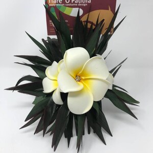 May include: White plumeria flower hair comb with a rhinestone in the center of the flower. The comb is surrounded by dark green leaves.