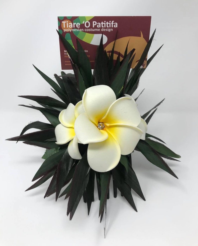 May include: White plumeria flower hair comb with a rhinestone in the center of the flower. The comb is surrounded by dark green leaves.
