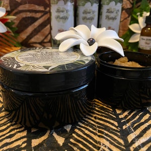 K&ouml;nnte beinhalten: Zwei schwarze Gl&auml;ser mit Deckeln, eines mit einem wei&szlig;en Etikett, auf dem "Tiare & Vanilla Sugar Scrub" steht, und das andere mit braunem Zucker gef&uuml;llt. Die Gl&auml;ser stehen auf einer braunen und schwarzen gemusterten Oberfl&auml;che.