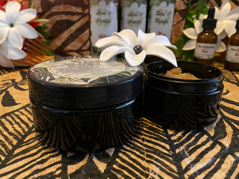K&ouml;nnte beinhalten: Zwei schwarze Gl&auml;ser mit Deckeln, eines mit einem wei&szlig;en Etikett, auf dem "Tiare & Vanilla Sugar Scrub" steht, und das andere mit braunem Zucker gef&uuml;llt. Die Gl&auml;ser stehen auf einer braunen und schwarzen gemusterten Oberfl&auml;che.