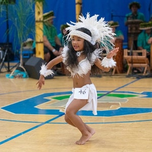 May include: A young girl in a white feathered headdress and costume performs a traditional dance on a wooden floor. She is wearing a white skirt with the number 46 on it.