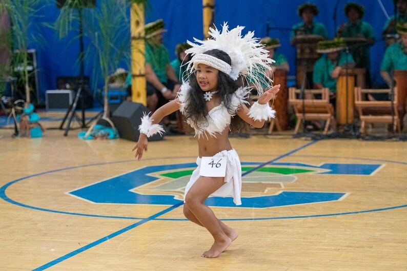 May include: A young girl in a white feathered headdress and costume performs a traditional dance on a wooden floor. She is wearing a white skirt with the number 46 on it.