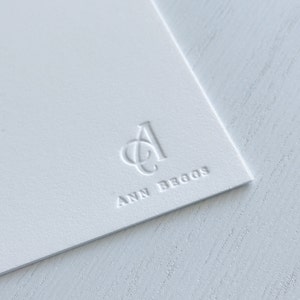 May include: A white card with a raised, embossed logo of the letters "A" and "C" intertwined. The text "ANN BEGGS" is embossed below the logo.