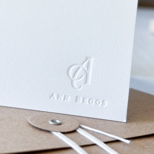 May include: A white card with a raised, embossed logo of the letters "AB" and the text "ANN BEGGOS" below the logo. The card is sitting on a brown envelope with white string.