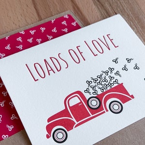 May include: A red pickup truck with a white bed filled with hearts. The truck has black wheels and a white cab. The text "LOADS OF LOVE" is printed in red on a white background.