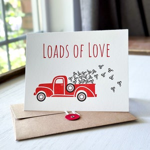 May include: A red vintage truck with a white bed is filled with black hearts. The truck is on a white card with the text "Loads of Love" in red.