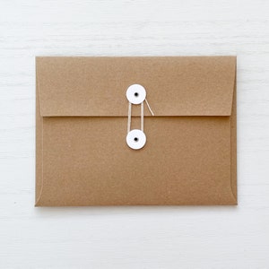May include: A brown paper envelope with a flap closure and a string tie. The envelope has two white circular fasteners with a string that wraps around them to close the envelope. The background is a white surface.