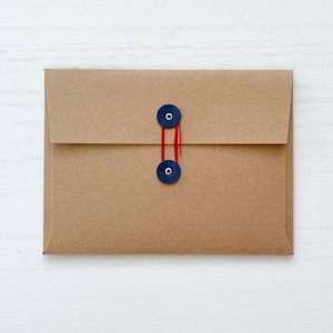 May include: A brown paper envelope with a flap closure. The envelope is secured with two navy blue buttons connected by a red string. The background is a white wooden surface.