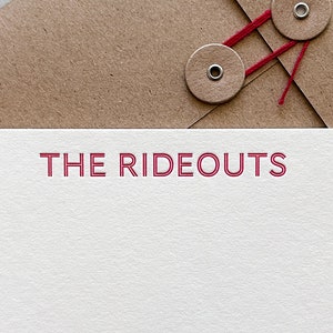 May include: A stationery set featuring a white card with the text "THE RIDEOUTS" in red, paired with a brown envelope. The envelope has two round, tan closures secured with red string.