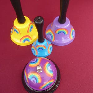 Op de afbeelding: Vier handbellen met regenboogontwerpen. De bellen zijn paars, blauw, geel en paars met zwarte handvatten.